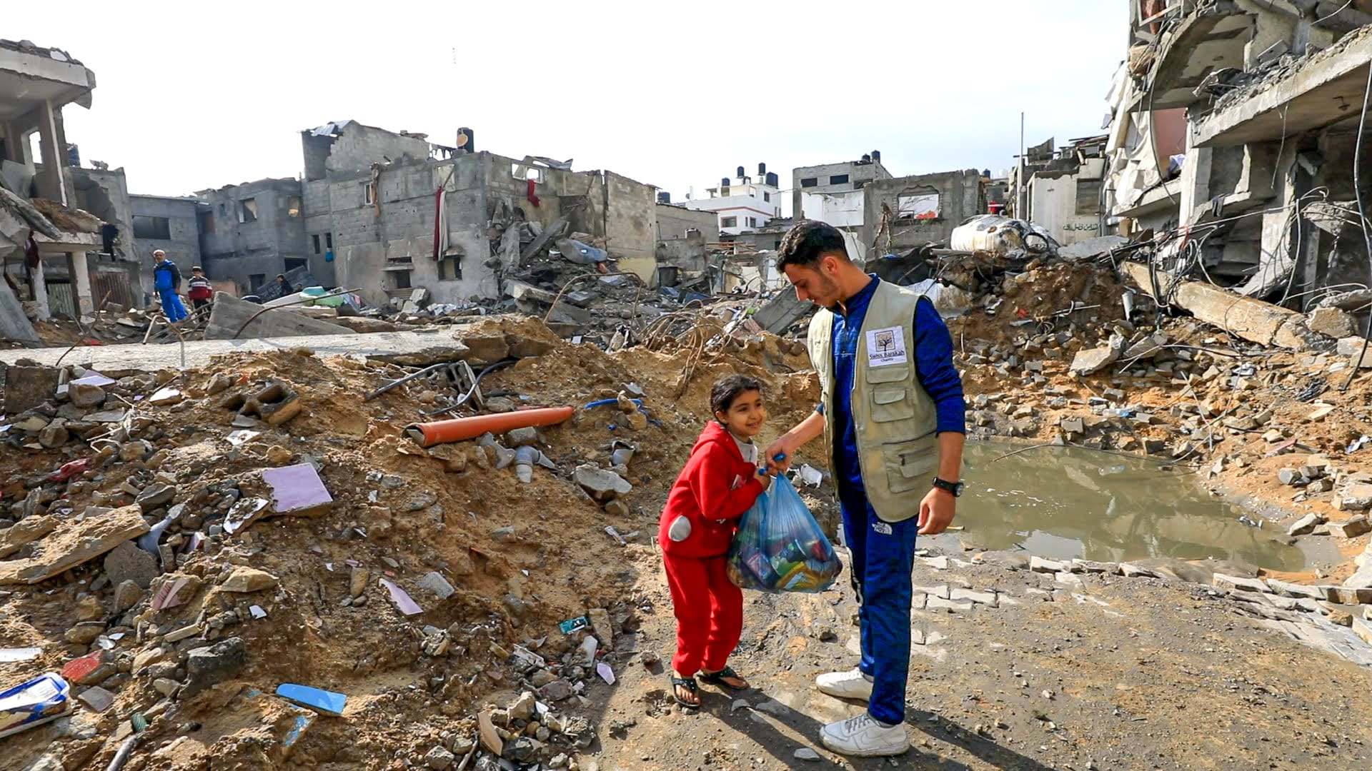 Un operatore umanitario consegna un sacchetto di cibo a una bambina sorridente in tuta rossa tra le macerie degli edifici.