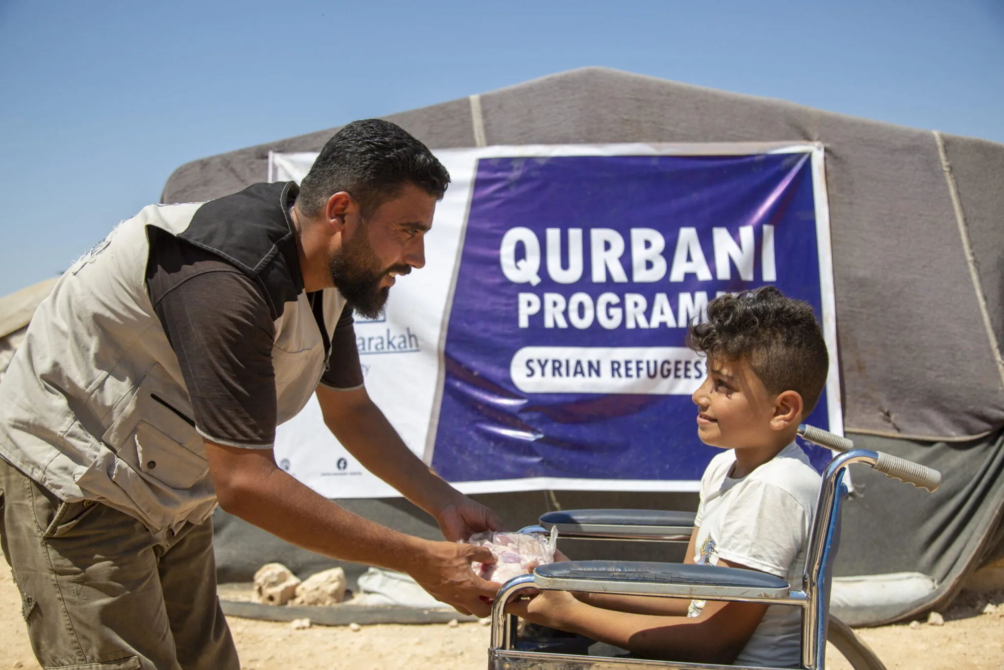 Ein Helfer überreicht einem Jungen im Rollstuhl ein Paket aus Kurban Spenden vor einem Hilfszelt.