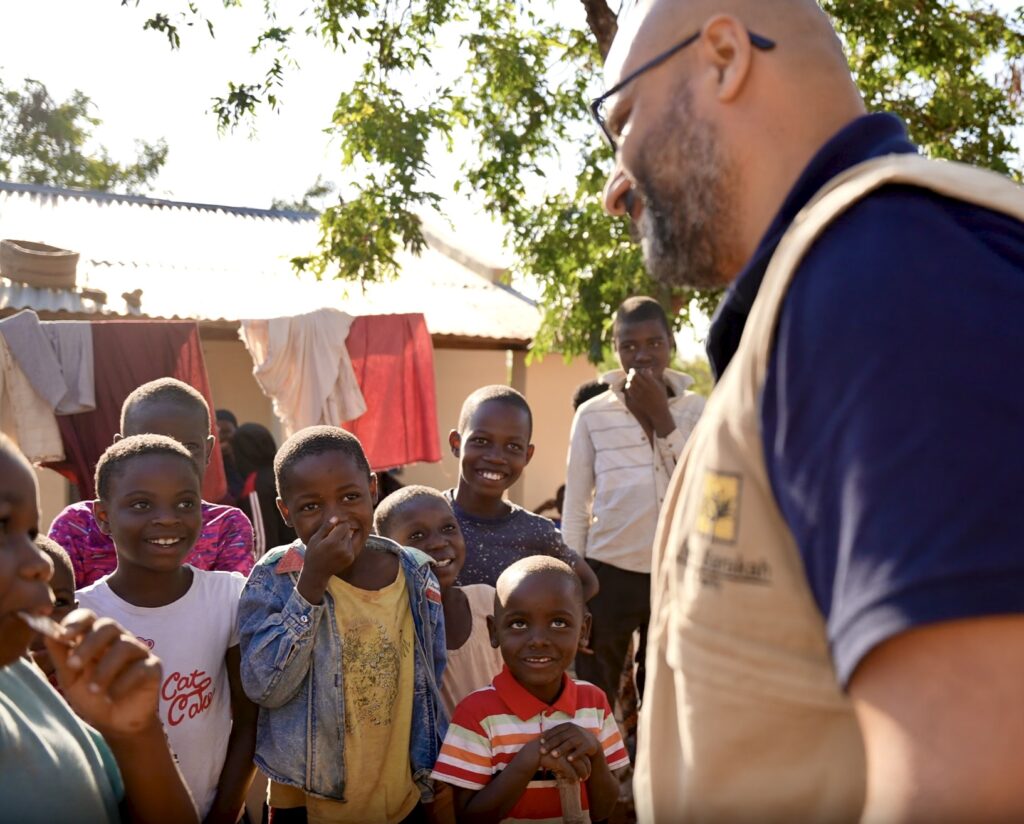 Ein lächelnder Entwicklungshelfer der Organisation Barakah spricht mit einer Gruppe Waisenkinder in einem afrikanischen Dorf.